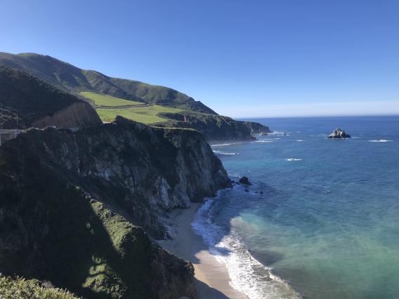The coast of California during midday with a cloudless blue sky and cliffs topped with greenery.