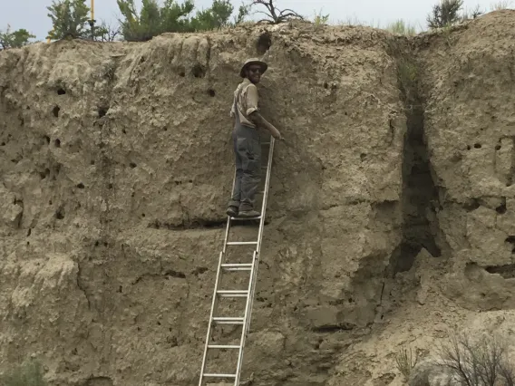 John Kemper standing on a ladder leaning on a cliffside looking back with a smile.