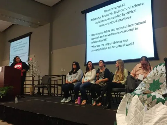 5 people sitting on a stage having an open discussion with a moderator off to the left.