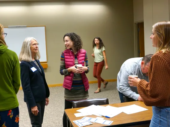 4 People stand around a table with documents on it, one person walking in the background