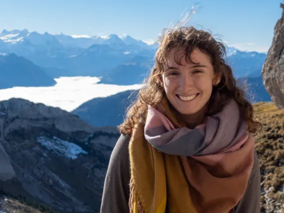 A young woman with curly brown hair is smiling at the camera while standing outdoors on a mountain trail. She is wearing a warm, multicolored scarf and a grey sweater. Behind her, there is a stunning backdrop of snow-capped mountains under a bright blue sky. The valley below is partially covered by a layer of clouds. The scene is bright and sunny, indicating a clear day in a high-altitude, mountainous area.