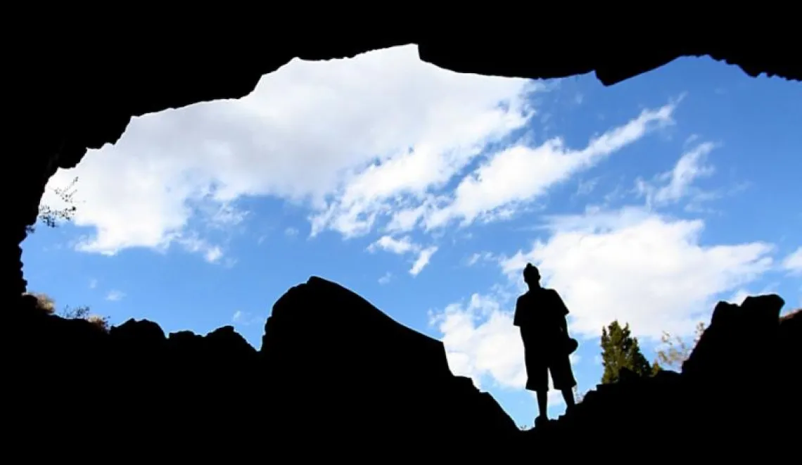 A silhouette of a man standing over an open crevice in the earth with a cloudy blue sky behind him.