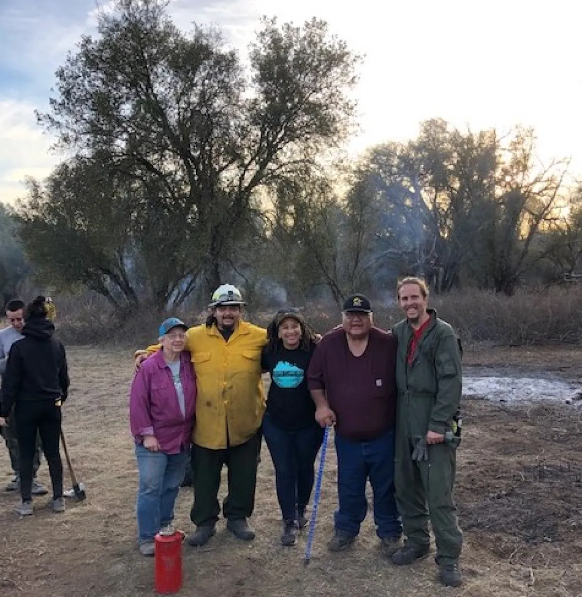 5 people standing next to each other with smoke from a controlled fire in the background.