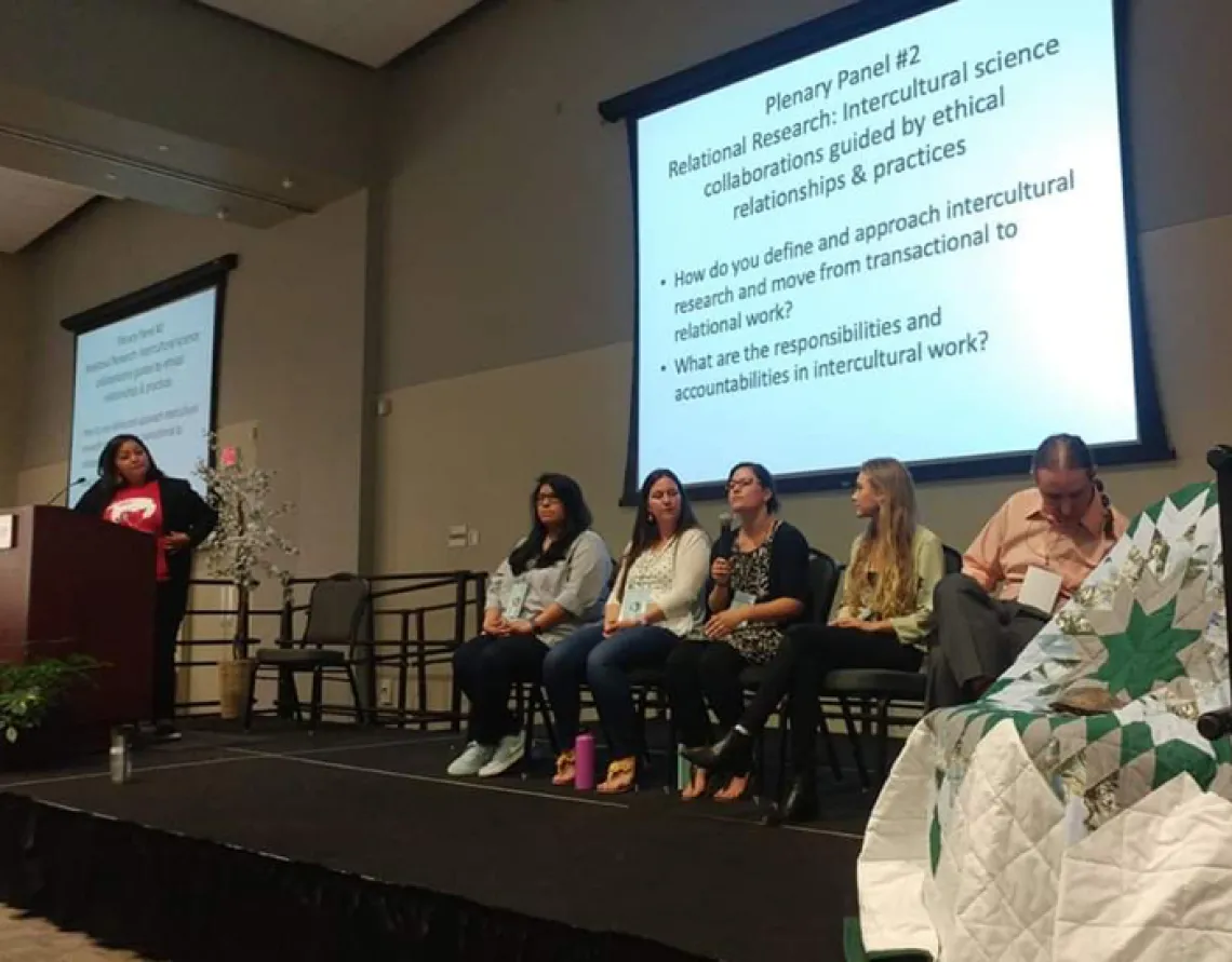 5 people sitting on a stage having an open discussion with a moderator off to the left.