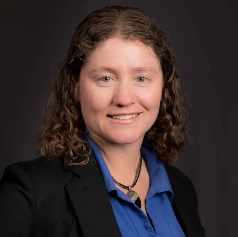 White woman with brown curly hair in a blue button up and suit jacket on a grey background