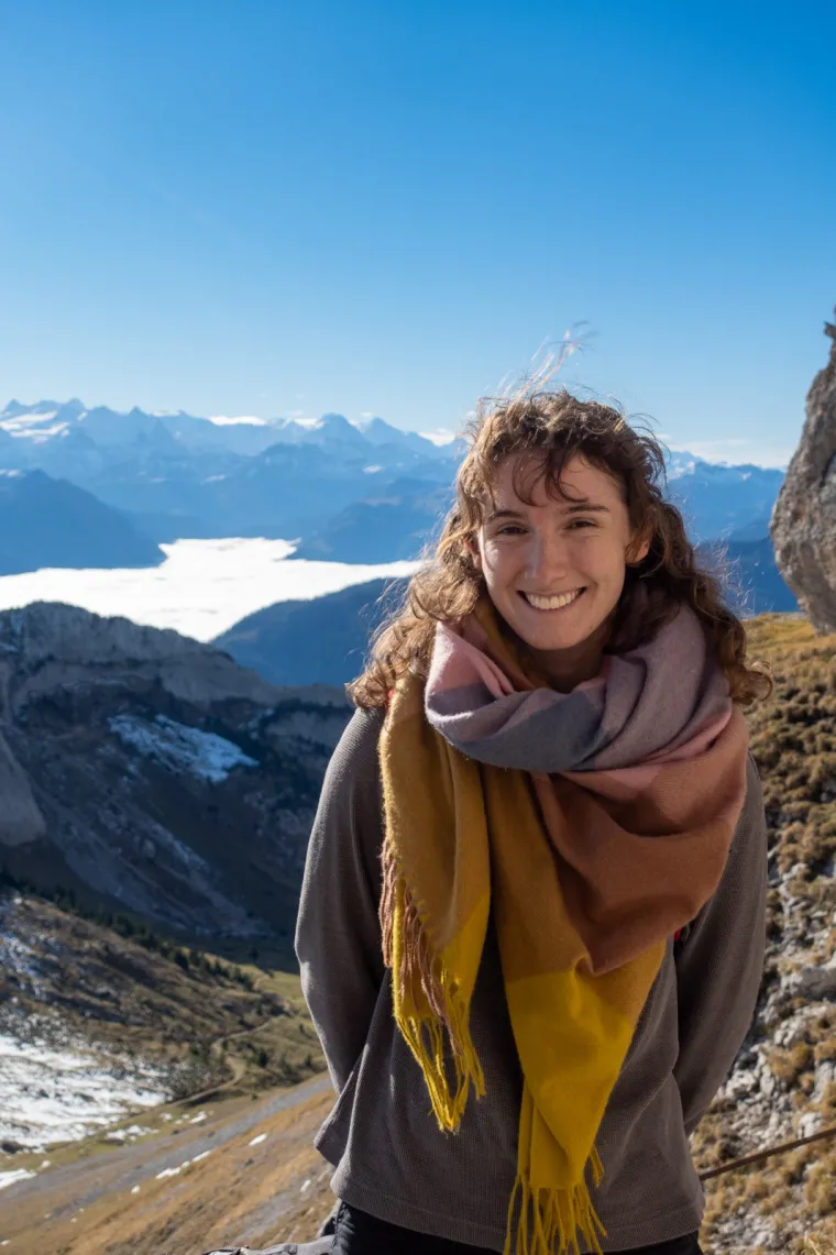 A young woman with curly brown hair is smiling at the camera while standing outdoors on a mountain trail. She is wearing a warm, multicolored scarf and a grey sweater. Behind her, there is a stunning backdrop of snow-capped mountains under a bright blue sky. The valley below is partially covered by a layer of clouds. The scene is bright and sunny, indicating a clear day in a high-altitude, mountainous area.