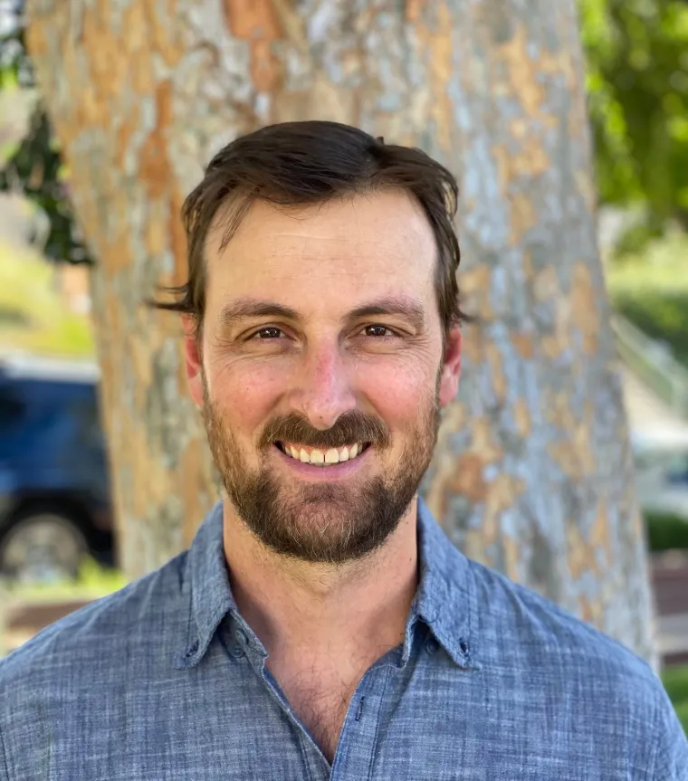 White man wiht short hair and beard in front of a tree wearing a button up grey shirt