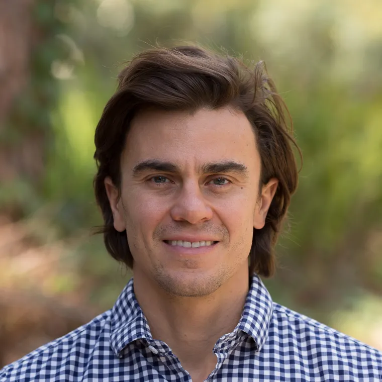 White man with medium short hair in plaid button up on blurry background of plants 