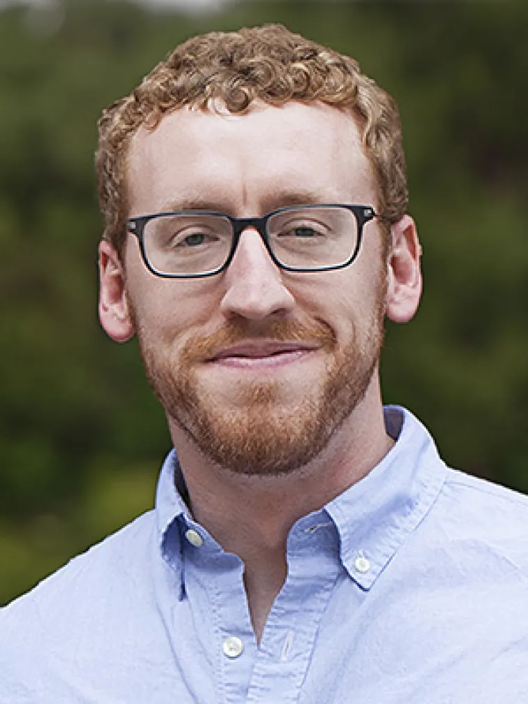 White man with short blonde hair wearing a light blue button up shirt on blurry green plant background